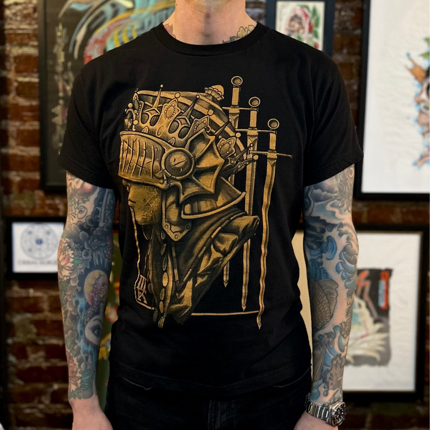 Person wearing a black t-shirt with a gold graphic design of a warrior women's helmet and swords, standing in front of tattoo studio walls.